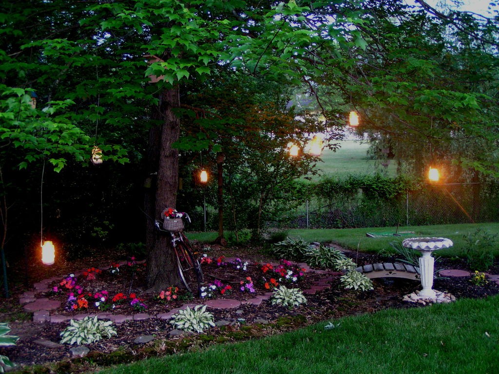 Yard surrounded with flowers and a big trees with candles in jars hanging on it