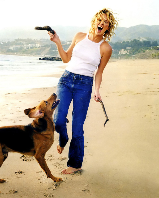 Woman in jeans and white top laughing and playing with a dog on the beach