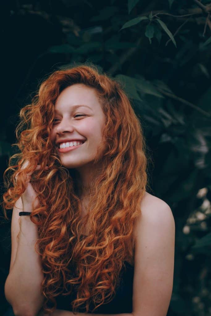Red-haired woman with light skin and curls smiling on a leafy background