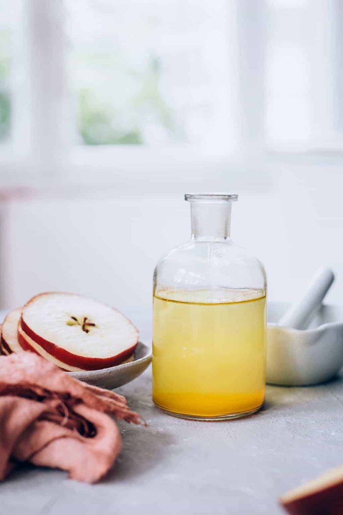 glass bottle of apple cider vinegar on a table with slices of apple in a plate