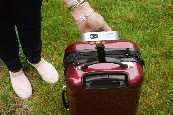 woman with a luggage scale weighing a red suitcase on a lawn