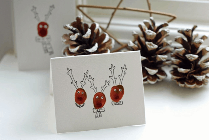 Fingerprint Reindeer DIY Christmas cards ideas white card with three reindeer pinecones in the background 