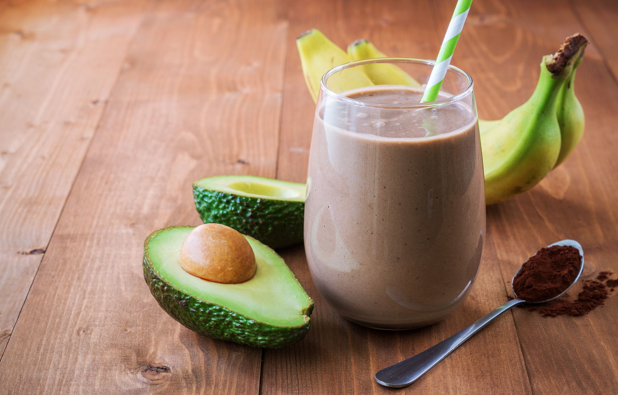 Chocolate protein in a glass with a straw next to banana and avocado