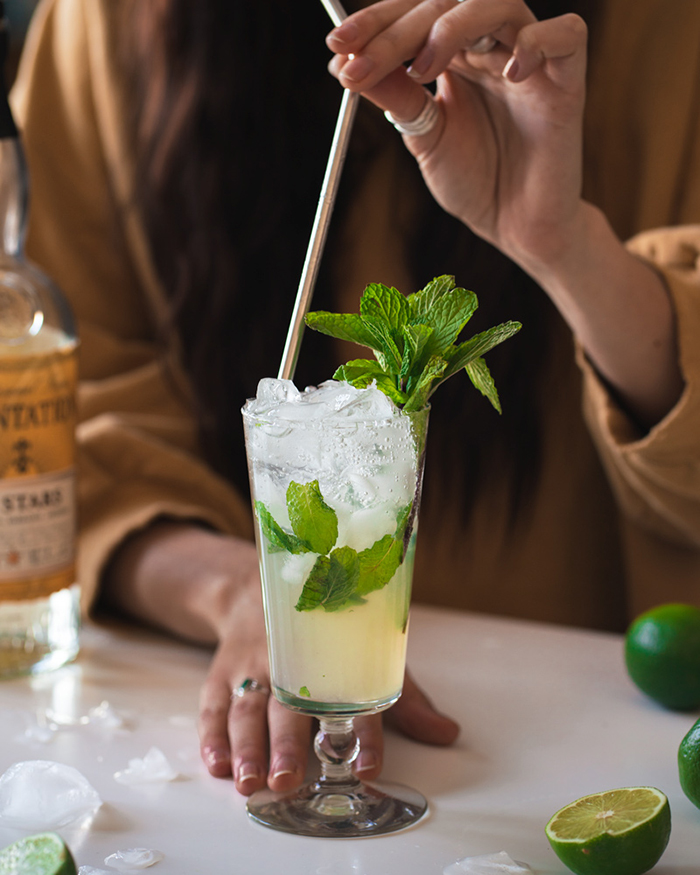 summer drinks ideas Mojito in a glass with ice and mint woman in the background holding a straw