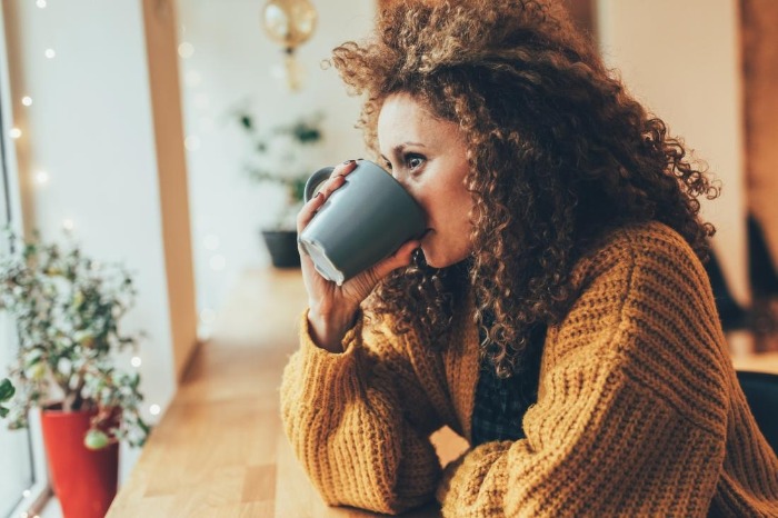 woman in a brown sweater with curly hair drinking from a grey mug in a cafe