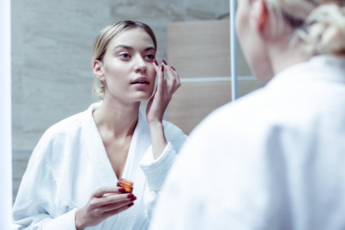 woman looking in the mirror applying hyaluronic acid on her face under her eyes 