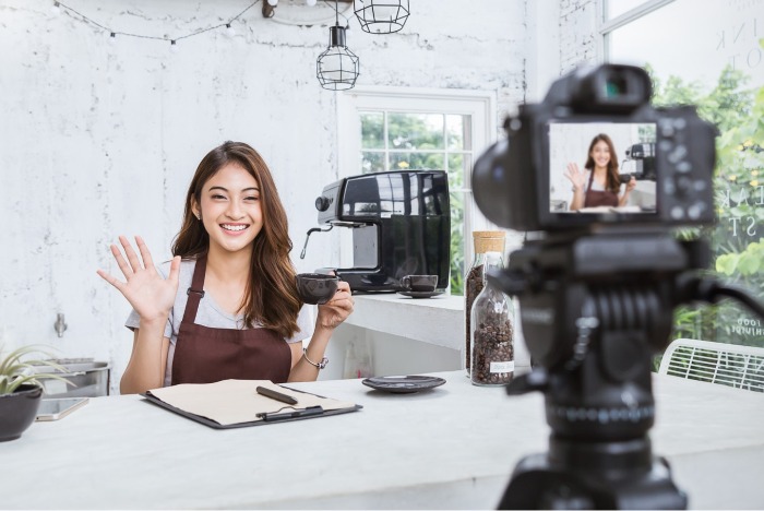 lifestyle influencer in apron waving at a camera set up with a cup of coffee 