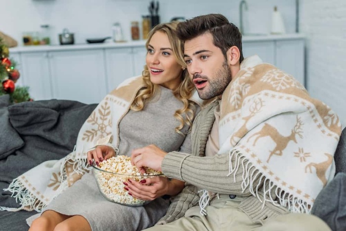 couple sitting on a sofa covered with a blanket watching christmas movies and eating popcorn 