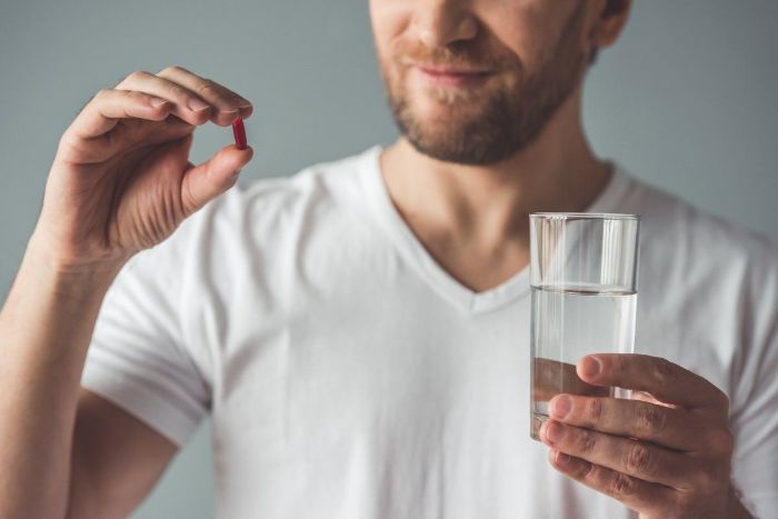 Man in white shirt with a beard holding a glass of water and a vitamin pill