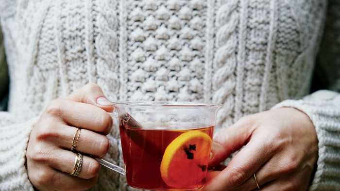 woman dressed in sweater holding a glass cup with warm tea cinnamon stick and orange peel