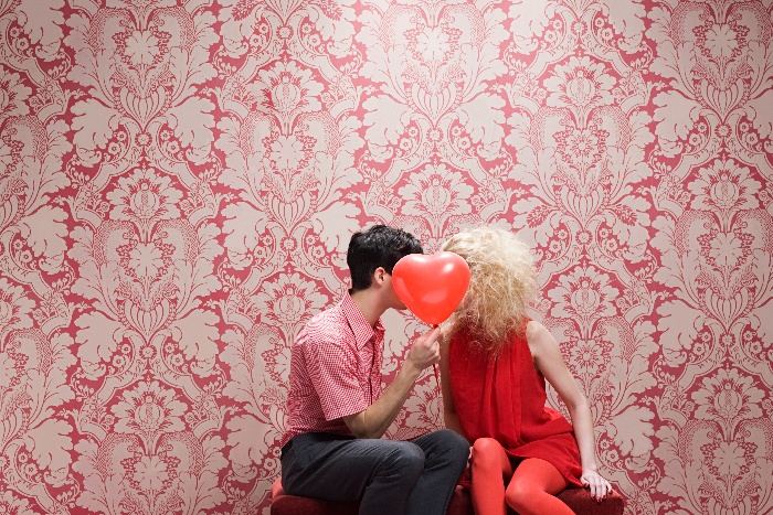 couple sitting on a bench kissing hinging behind a heart-shaped red balloon