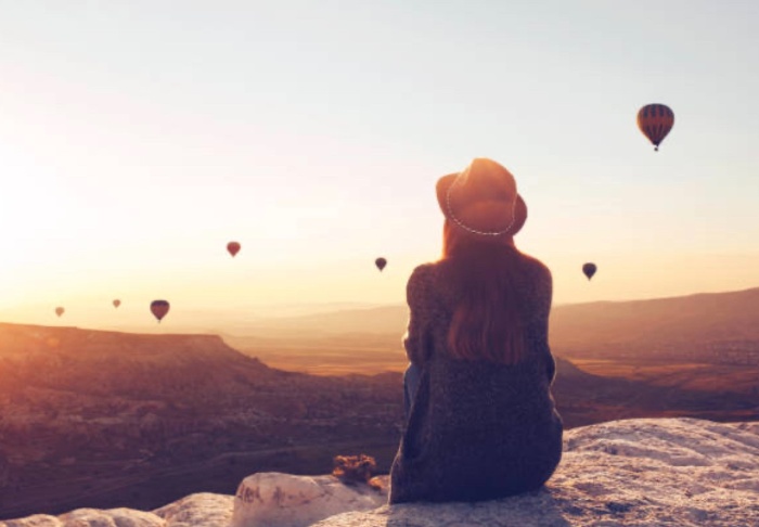 woman sitting on a rock daydreaming watching hot air balloons in the horizon