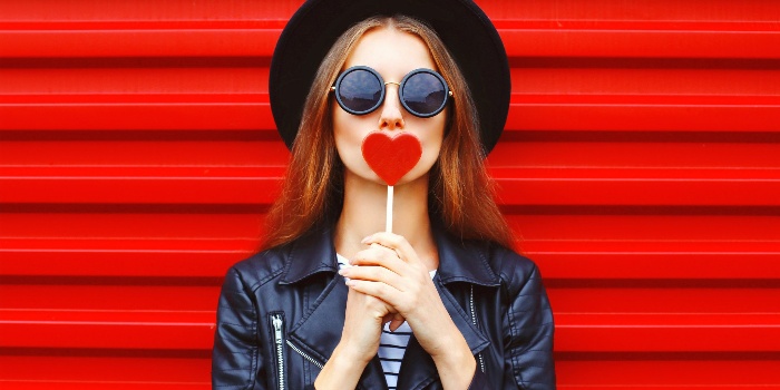 love archetype woman with black sunglasses hat and leather jacket on a red background holding a heart in her hands