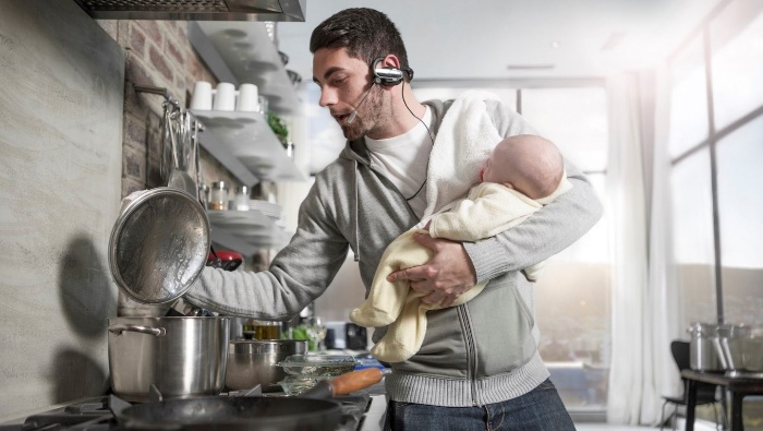 new family roles a man multitasking holding a baby cooking and speaking on a hands free