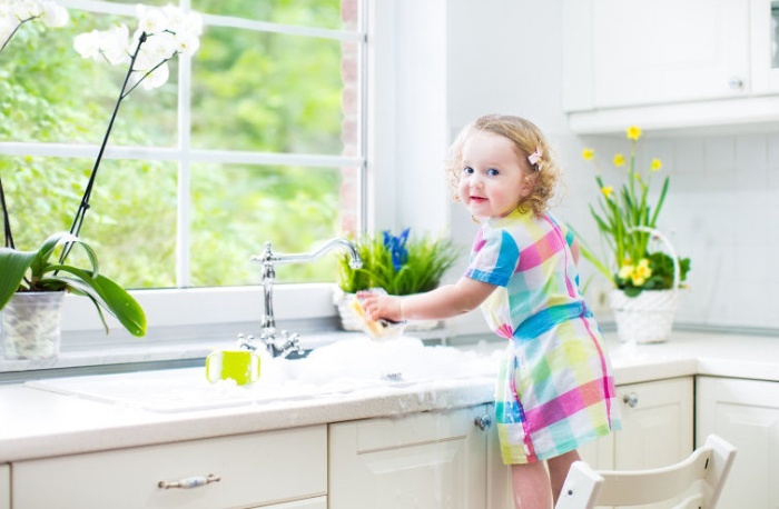 little girl in a colorful dress standing on a chair and cleaning a white kitchen with some plants