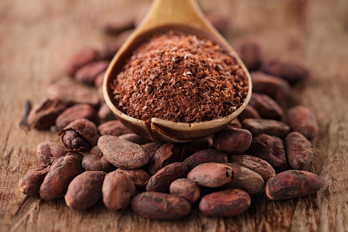 wooden spoon with cocoa powder on a wooden table and a pile cocoa beans