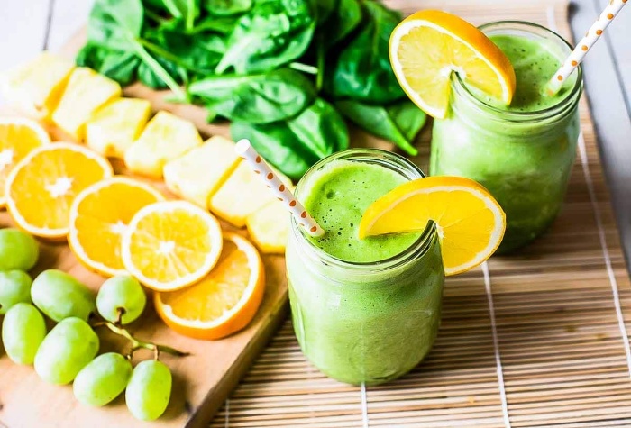 healthy juices green juices in jars with straws and lemon slices in front of a cutting board with different fruits and spinach