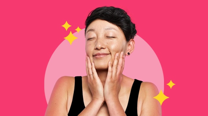 woman with black hair and black top enjoying her healthy and glowing skin on a pink background with her eyes closed