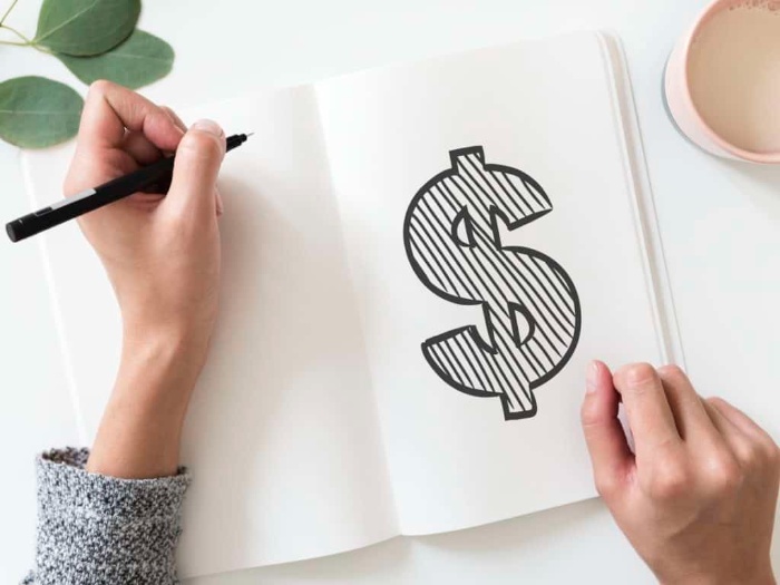 money saving tips woman writing a dollar sign into a white notebook with a black pen