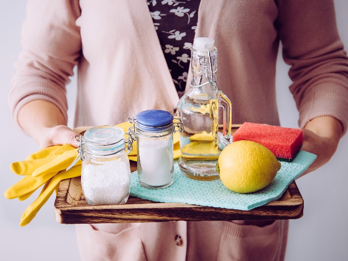 woman in pink cardigan holding a tray with natural cleaning products soda lemon yellow gloves