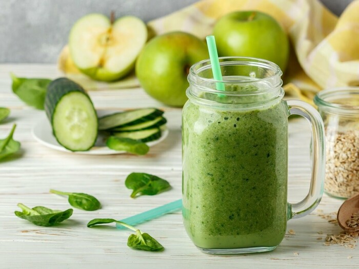 cucumber juice in a glass jar with a green straw on a white table with green apples cucumbers and mind leaves