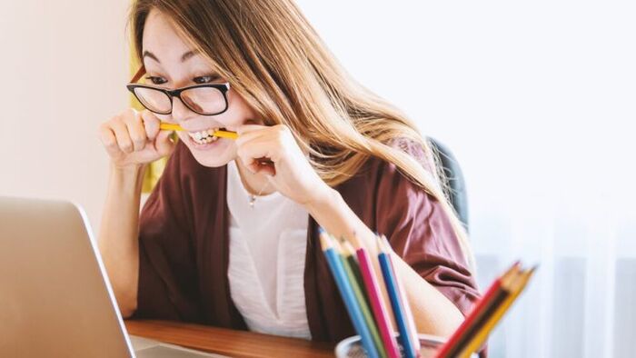 asian woman with glasses and long hair looking at her pc with a pen in her mouth