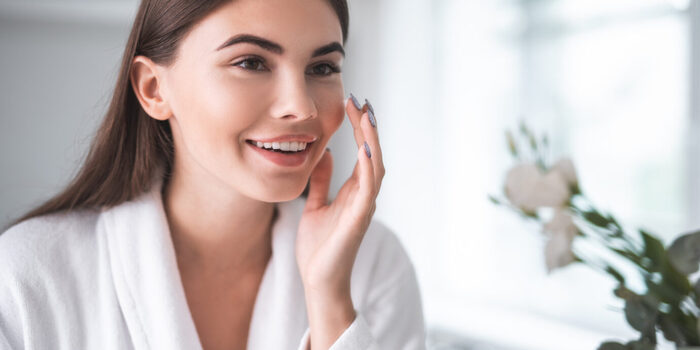 Portrait of pleased young lady in a white bathrobe touching her cheek with hand putting cream on her face skin indoor