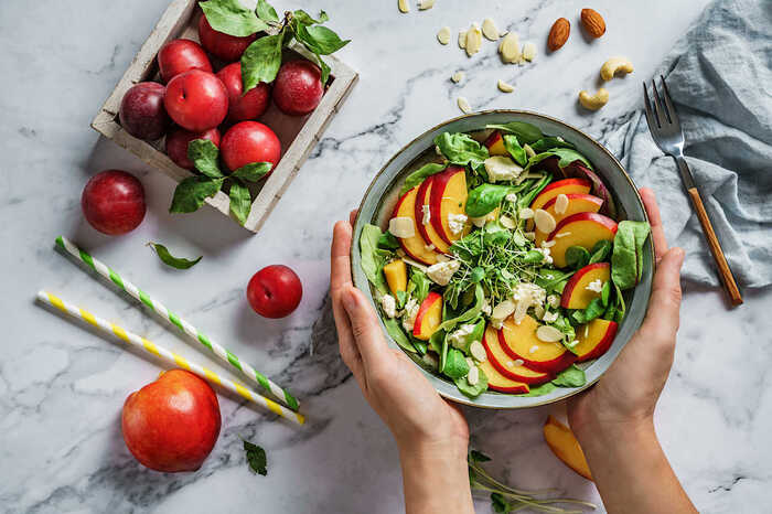 eat fruits woman holding a bowl with salad and peach slices inside on a marble table with other fruits around