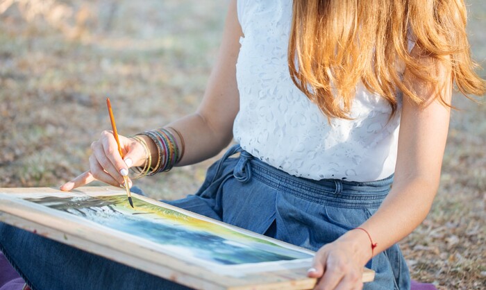 woman with long light hair in denim skirt and white top sitting outdoors painting 