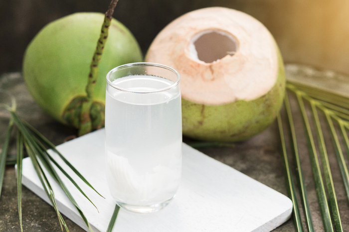 fresh coconut water full of minerals in a tall glass on a white plate with two open green coconuts in the background and palm leaves