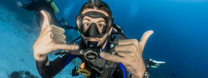 beginner scuba diver man in his diving costume showing a finger sign and looking into the camera deep underwater