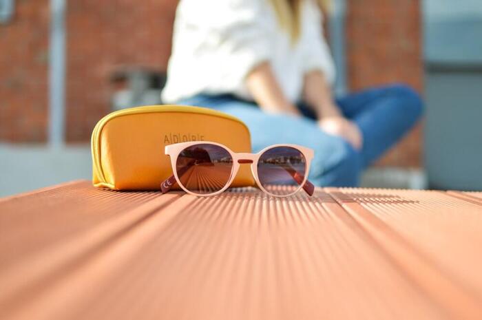 branded sunglasses in light pink on a table next to a mustard pouch case with a woman in white shirt and jeans in the background