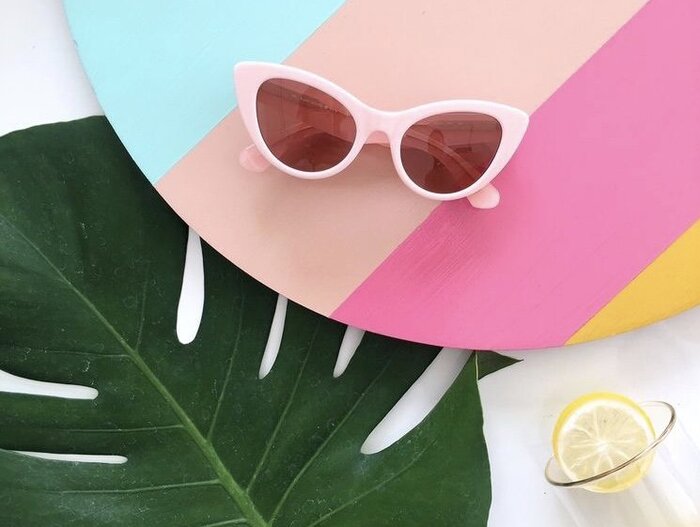 cat eye sunglasses in baby pink on a colorful surface with a large green leaf next to them and a glass with a lemon slice