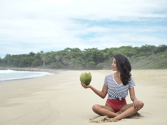 woman with dark hair and striped shirt sitting in a yoga pose on a wide breach holding a large green coconut in her hand