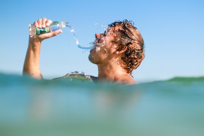 blond man swimming in the sea squeezing a water bottle out at his face