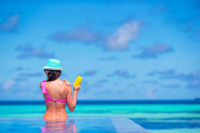 woman in a pool with pink swimsuit and bright blue hat applying sunscreen on her shoulder