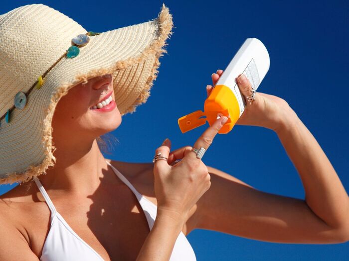 woman with a sun hat and white swim suit smiling and applying sun lotion from a bottle