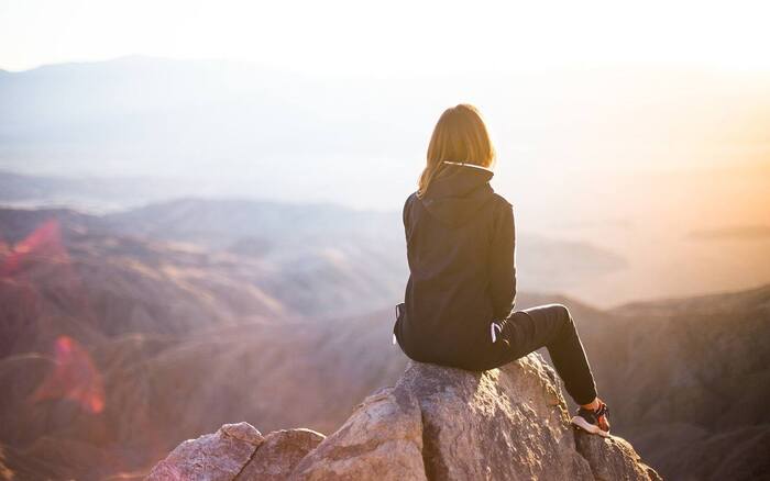 woman dressed in jacket and pants solo traveler sitting on a rock overlooking a hillside landscape at sunset