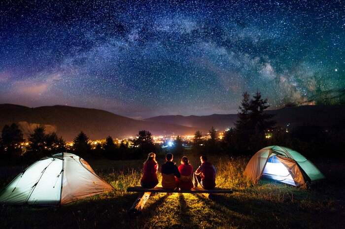 summer camping family sitting on a wooden bench between two tents overlooking the starry night around a fire