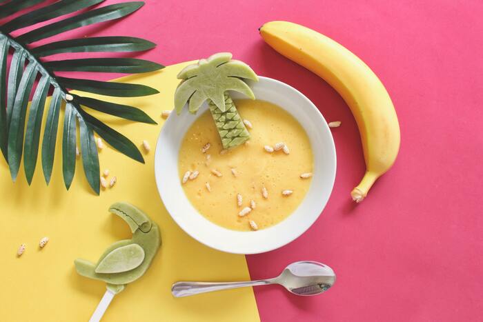 sweet fun food a white bowl full of yellow smoothie with a palm leaf banana tea spoon and decorations