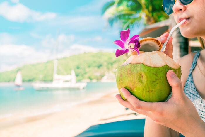 woman holding and drinking from a green coconut on a beach with a little purple flower on the coconut