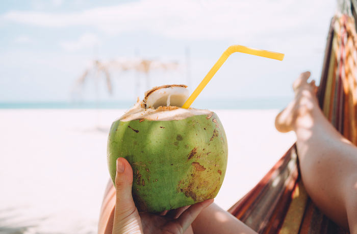 Woman lying on the beach holding a green coconut with a yellow straw in it relaxing and drinking coconut water