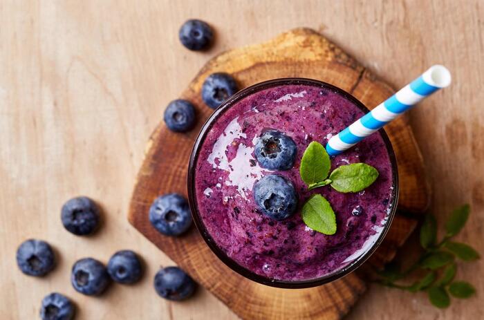 blueberry smoothie with blue and white straw and blueberries scattered around on a wooden piece