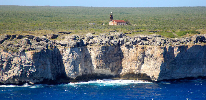 isla de mona with high cliffs and blue waves crashing in them with a lighthouse on the island