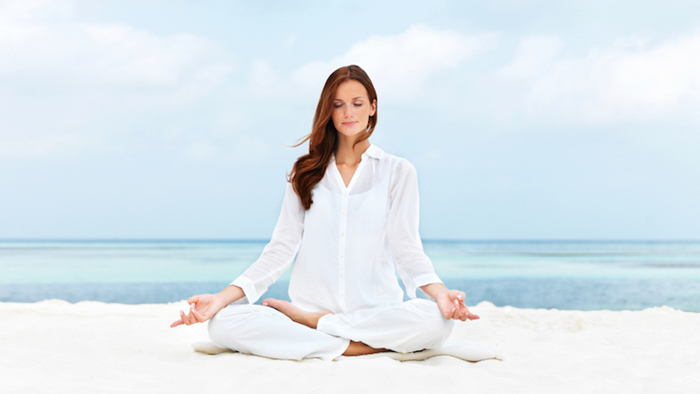 woman sitting on a white sand beach and dressed in white meditating in a lotus pose