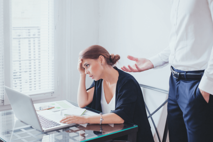 narcissistic boss woman sitting in front of a computer holding her forehead and looking annoyed while a man is standing beside her explaining something