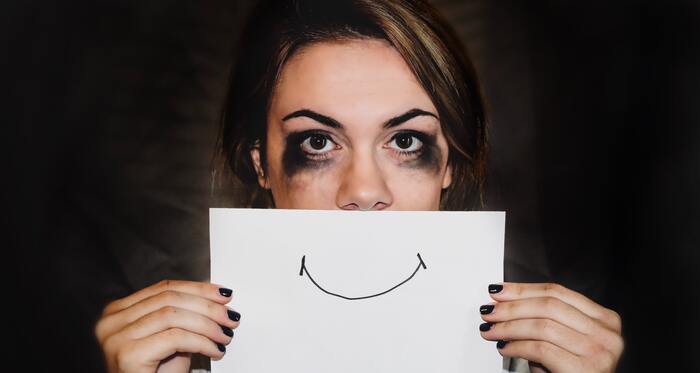 social anxiety woman crying with smudged make up holding a white piece of paper with a smile in front of her face