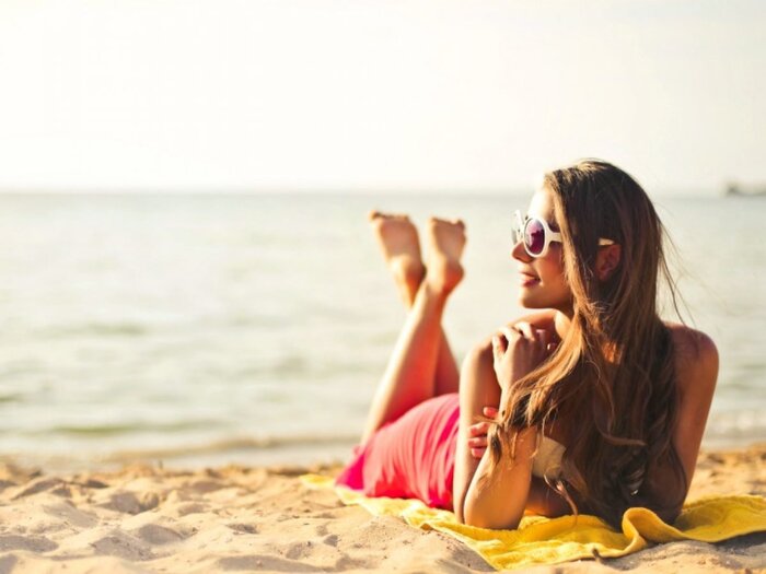sunbathing woman in a pink dress with white sunglasses and log hair lying on the beach on a yellow towel lookin in one direction