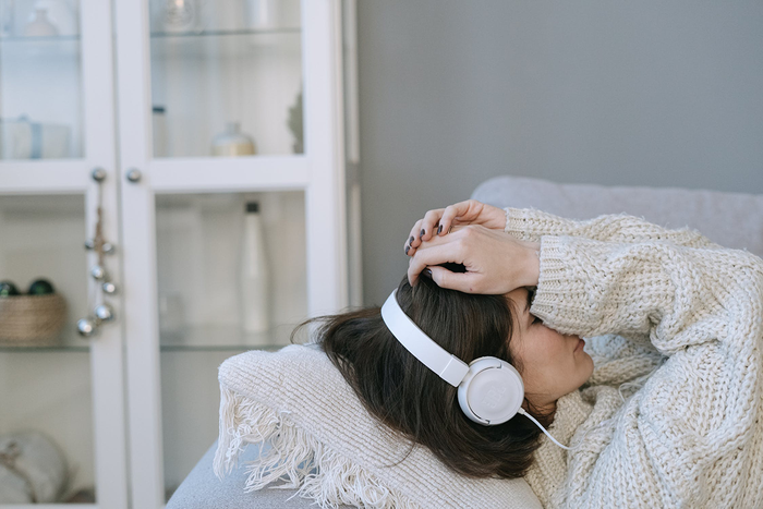 woman in a chunky white sweater listening to music relaxing on a couch and holding her head with her hands