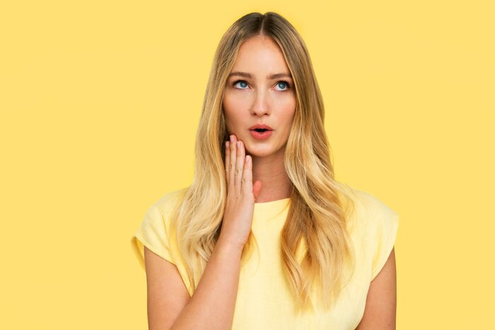 blond haired woman dressed in a light yellow blouse on a yellow background looking surprised touching her face with a hand